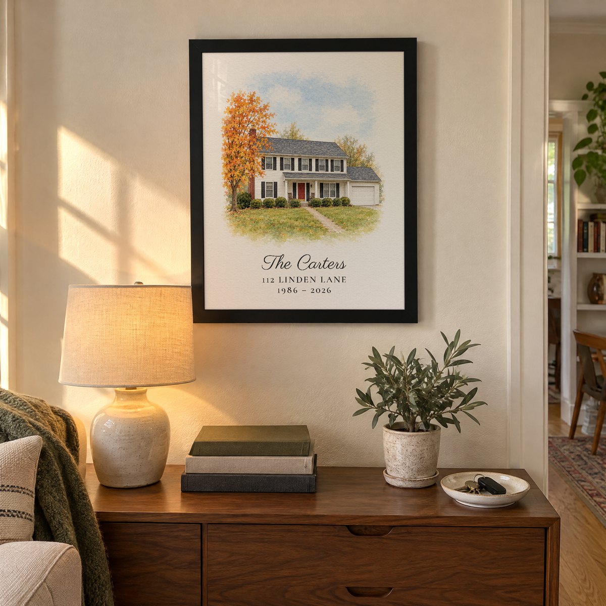 Custom house portrait of a white colonial with a red door, in a black frame above a living-room console, with the line The Carters, 112 Linden Lane, 1986 - 2026