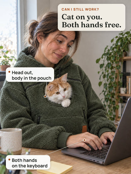 A woman types with both hands while her orange-and-white cat watches from the hoodie's round chest opening