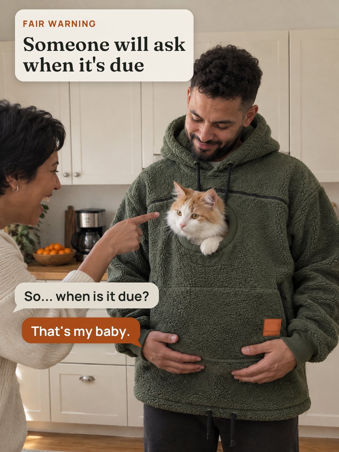 A friend laughs and points at the orange-and-white cat peeking out of a man's hoodie in the kitchen