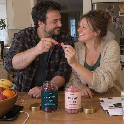 A couple at their kitchen table doing a playful cheers with their gummies, the HIM and HER jars in front of them