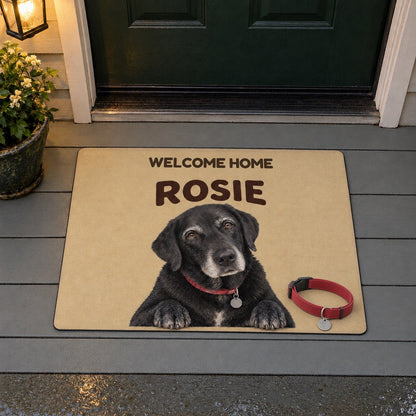 A doormat with a black Labrador named Rosie and the words Welcome home, her red collar resting on it, on a porch at dusk