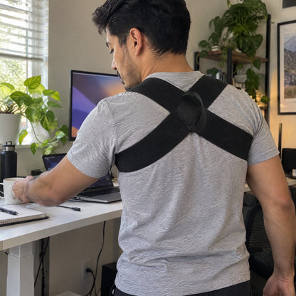 Man at a standing desk wearing the black X-back posture strap over a grey T-shirt