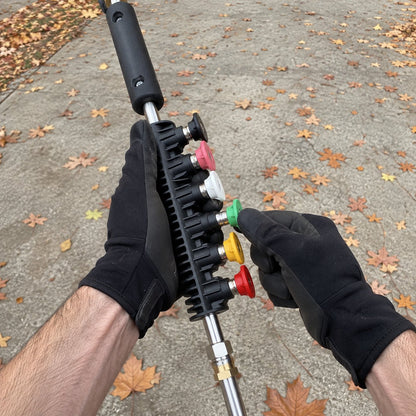 Gloved hands pulling the green spray tip out of the black tip holder on the handle, next to the black, pink, white, yellow and red tips