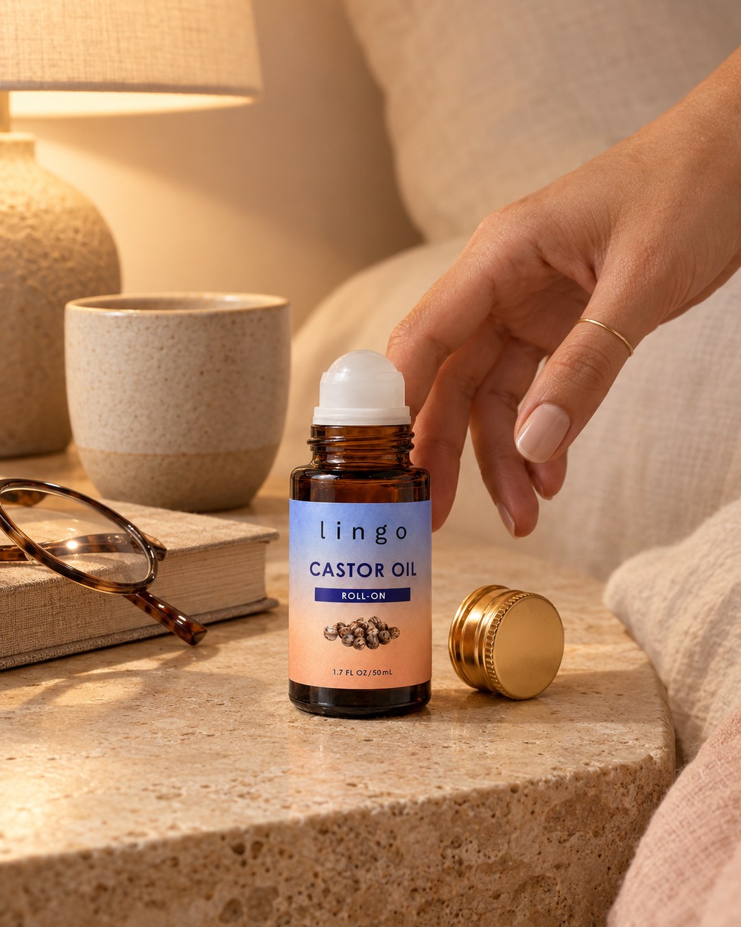 The castor oil roll-on on a stone nightstand next to reading glasses and a book, a hand reaching for it