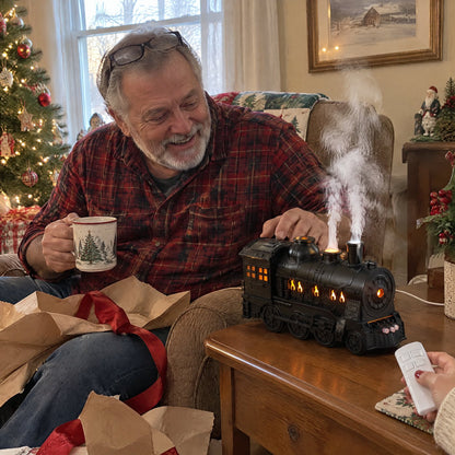 A man smiling at his new steam train diffuser puffing mist beside his armchair on Christmas morning