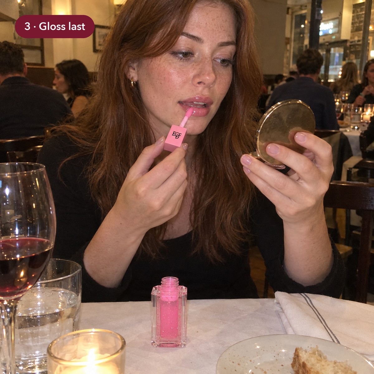 Woman applying pink lip gloss with a compact mirror at a restaurant table, the gloss tube in front of her