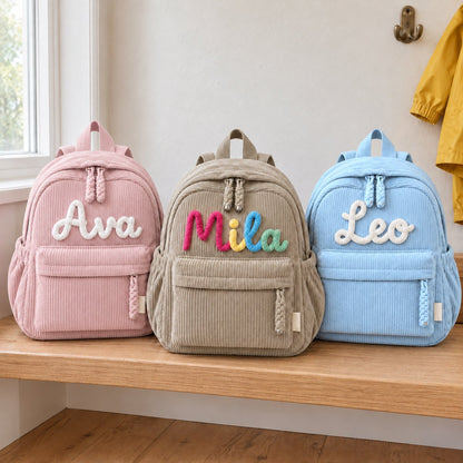 Three kids' corduroy backpacks on a window bench: pink with Ava, khaki with Mila in rainbow letters and blue with Leo