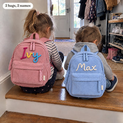 Two siblings on a step seen from behind, each with their own named backpack: pink Ivy and blue Max